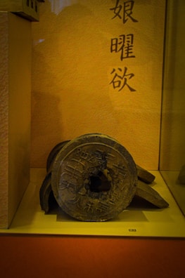 An ancient, round, metal object with inscriptions rests on a display stand. The background consists of a warm, yellowish wall with vertical Asian script.