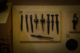 A collection of ancient black metal tools and artifacts displayed on a beige board, each with distinct shapes and sizes. The items appear to be old and possibly handcrafted, arranged neatly in a museum-like setting with numerical labels.