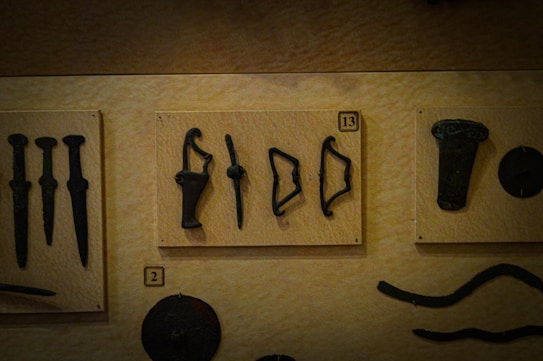 A collection of ancient artifacts is displayed on a beige background. The artifacts include various tools and weapons. The central panel, labeled number 13, holds five distinct items including hooks and unidentified tools. The left panel shows three elongated objects resembling daggers, marked with the number 2. An assortment of dark, metallic objects, including possibly a shield or plaque, is on the right and lower sections.