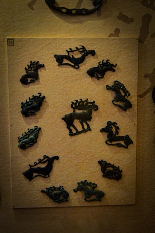 A display of twelve black metal or stone animal-shaped artifacts is mounted on a light-colored board. The artifacts resemble various animals such as deer and other quadrupeds with curved and intricate designs. The board is secured to a wall decorated in a subtle, textured pattern. The overall lighting is low and focused primarily on the artifacts.