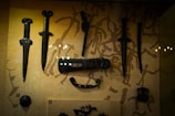 A collection of ornate, ancient-looking daggers and other weapons displayed on a background with a pattern of abstract figures. The weapons are dark, possibly made of metal, and have intricate designs, showcasing historical craftsmanship.