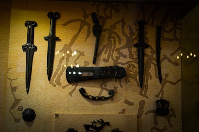 A collection of ornate, ancient-looking daggers and other weapons displayed on a background with a pattern of abstract figures. The weapons are dark, possibly made of metal, and have intricate designs, showcasing historical craftsmanship.