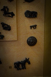 Various dark, metal figurines of animals are displayed on a textured beige background. The figurines include an elephant, a donkey, and some other indistinct shapes. A decorative circular metal piece is also present.