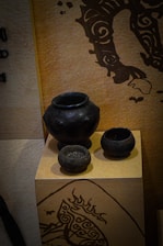 Several black pottery vessels are displayed on a beige pedestal against a textured background with artistic black patterns. The vessels vary in size and shape, featuring a matte finish. The background design includes swirling motifs that add a decorative element to the setting.