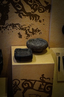 Two stone artifacts are displayed on a pedestal, with intricate ancient patterns and motifs decorating the background wall. The stones have smooth surfaces, with one being more rounded and the other more rectangular. The setting gives a sense of historical or archaeological significance, possibly from a museum exhibit.