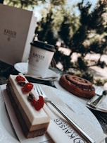 A close-up view of a dessert table setup featuring a layered cake topped with raspberries and pistachios, alongside a cinnamon roll. A cup with a lid and napkins are also visible, branded with 'Kaiser Patisserie', set on a plate and a wooden surface. The background shows defocused greenery, suggesting an outdoor setting.