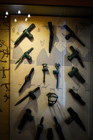 A display case containing a collection of ancient bronze tools and weapons, including axes, daggers, and hammers. The background features silhouettes of figures and animals, resembling cave art.
