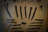 A collection of ancient metal tools and weapons displayed inside a glass case. The items include various knives, daggers, and possibly hairpins or similar objects with intricate designs. The background is a textured beige surface that enhances the visibility of the dark tools.