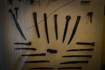 A collection of ancient metal tools and weapons displayed inside a glass case. The items include various knives, daggers, and possibly hairpins or similar objects with intricate designs. The background is a textured beige surface that enhances the visibility of the dark tools.