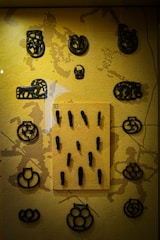 A collection of small, intricately designed black items displayed on a textured yellow background. The items appear to be made of metal and have various shapes and artistic patterns. Some are circular with symmetrical designs, while others have more abstract forms. A central rectangular panel holds several small, elongated objects.