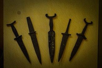 A collection of five ancient metal daggers displayed on a flat, light-colored surface. Each dagger has a distinct hilt design, with some featuring simple crossguards and others having more elaborate forms. The blades appear aged and are dark in color, suggesting they are made from iron or a similar material.