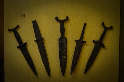 A collection of five ancient metal daggers displayed on a flat, light-colored surface. Each dagger has a distinct hilt design, with some featuring simple crossguards and others having more elaborate forms. The blades appear aged and are dark in color, suggesting they are made from iron or a similar material.