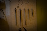 A set of five metal tools with a pointed end displayed vertically against a beige background. Each tool varies in length and design, with some featuring loops or rings at the top. There is a small numbered label '11' placed above the second tool from the left.