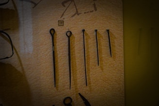 A set of five metal tools with a pointed end displayed vertically against a beige background. Each tool varies in length and design, with some featuring loops or rings at the top. There is a small numbered label '11' placed above the second tool from the left.