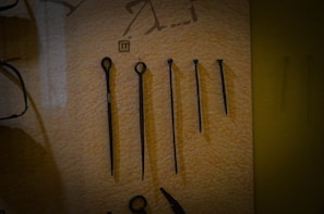 A set of five metal tools with a pointed end displayed vertically against a beige background. Each tool varies in length and design, with some featuring loops or rings at the top. There is a small numbered label '11' placed above the second tool from the left.