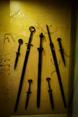 A collection of ancient swords and daggers displayed against a backdrop featuring primitive cave drawings. The weapons vary in size, with intricate designs on the hilts. The background adds historical context with sketches of animals and figures.