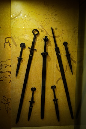 A collection of ancient swords and daggers displayed against a backdrop featuring primitive cave drawings. The weapons vary in size, with intricate designs on the hilts. The background adds historical context with sketches of animals and figures.