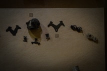 Various ancient artifacts displayed on a textured surface. The items include small metallic objects with intricate designs, possibly made of bronze or iron. Some objects are triangular, others are more abstract in shape, and there are numbers indicating their catalog information.