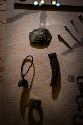 Various ancient metal artifacts are displayed against a textured backdrop. The collection includes small tool-like objects, possibly blades or chisels, a curved piece with a loop, and a rectangular item with engraved patterns.