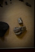 A display case contains ancient artifacts set against a textured, beige background. Among these objects, there is a prominently visible sculpted piece resembling a face fragment, emphasizing the lips and nose. There are additional smaller artifacts placed above, and a larger dark object to the left.
