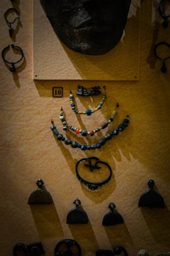 A display of various historical artifacts mounted on a textured wall. At the top is a face mask or sculpture made of a dark material. Below it are several strands of beaded necklaces composed of small colorful beads with diverse shapes. Surrounding these are several small metallic ornaments, possibly ancient pendants or tools, displayed in an organized manner.
