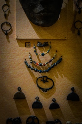 A display of various historical artifacts mounted on a textured wall. At the top is a face mask or sculpture made of a dark material. Below it are several strands of beaded necklaces composed of small colorful beads with diverse shapes. Surrounding these are several small metallic ornaments, possibly ancient pendants or tools, displayed in an organized manner.
