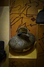 A dark, intricately patterned ceramic vessel displayed against a backdrop featuring abstract, linear art. The vessel, possibly historical or cultural, sits on a light brown platform, highlighting its textured surface. The background art is in dark brown lines on an earthy, tan surface.
