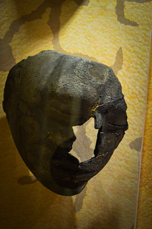 Close-up of a cracked, bone-white papier-mâché mask with haunting hollow eyes and jagged edges.