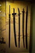 Several ancient swords are displayed upright against a backdrop with prehistoric art. The swords vary slightly in size and shape, and numbered labels are attached to each one. The background features abstract patterns and symbols, hinting at historical or cultural significance.