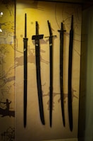 Several ancient swords are displayed upright against a backdrop with prehistoric art. The swords vary slightly in size and shape, and numbered labels are attached to each one. The background features abstract patterns and symbols, hinting at historical or cultural significance.