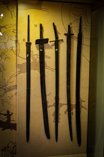 Several ancient swords are displayed upright against a backdrop with prehistoric art. The swords vary slightly in size and shape, and numbered labels are attached to each one. The background features abstract patterns and symbols, hinting at historical or cultural significance.