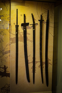 Several ancient swords are displayed upright against a backdrop with prehistoric art. The swords vary slightly in size and shape, and numbered labels are attached to each one. The background features abstract patterns and symbols, hinting at historical or cultural significance.