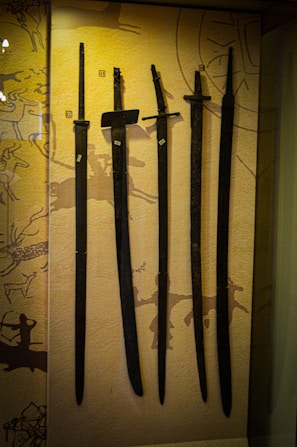 Several ancient swords are displayed upright against a backdrop with prehistoric art. The swords vary slightly in size and shape, and numbered labels are attached to each one. The background features abstract patterns and symbols, hinting at historical or cultural significance.
