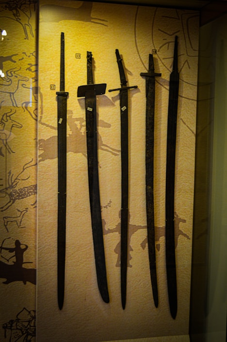 Several ancient swords are displayed upright against a backdrop with prehistoric art. The swords vary slightly in size and shape, and numbered labels are attached to each one. The background features abstract patterns and symbols, hinting at historical or cultural significance.