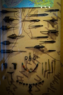 A display case contains an assortment of historical artifacts, including various spearheads and arrowheads arranged neatly. There is a map on the upper section, while below are several necklaces and implements. Each item is labeled with a number for identification. The background is textured, likely representing ancient artwork or symbols.