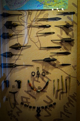 A display case contains an assortment of historical artifacts, including various spearheads and arrowheads arranged neatly. There is a map on the upper section, while below are several necklaces and implements. Each item is labeled with a number for identification. The background is textured, likely representing ancient artwork or symbols.
