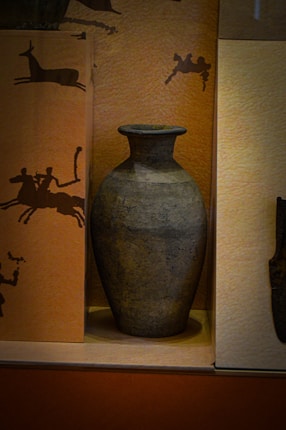 A large, ancient-looking ceramic vase is showcased against a background adorned with silhouettes of animals and human figures in various activities. The vase has a wide body and a narrow neck, with a dark, textured surface indicating age or crafting style.