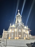 A well-lit church building at night with a prominent clock tower, multiple arched windows, and a cross on top. The building is illuminated by lights casting beams into the sky. Stairs lead up to the entrance with a person standing by the door. A large cross is present to the right of the church.