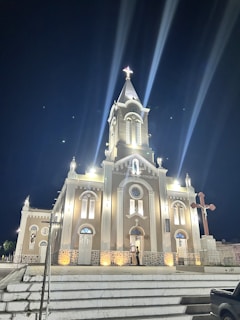 A well-lit church building at night with a prominent clock tower, multiple arched windows, and a cross on top. The building is illuminated by lights casting beams into the sky. Stairs lead up to the entrance with a person standing by the door. A large cross is present to the right of the church.