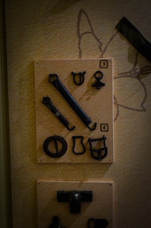 A display of several metallic, antique objects mounted on a textured background. The objects vary in shape and size, appearing to be hooks, rings, and other tools or hardware. They are placed in two separate panels, each marked with numbers '3' and '4'.