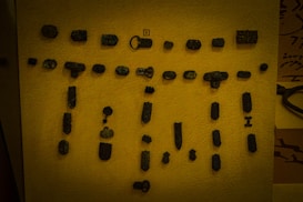 A collection of small, ancient metal artifacts displayed on a flat surface. The objects include various shapes and sizes, such as keys, intricate designs, and possibly fragments of tools or decorative pieces. The background is a muted yellow, contrasting with the dark color of the artifacts.