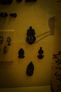 A collection of dark ancient artifacts displayed on a textured yellow background. The objects appear to be made of metal and include various intricate designs, some resembling abstract shapes and others more ornate with detailed carvings.