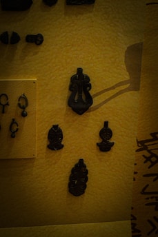 A collection of dark ancient artifacts displayed on a textured yellow background. The objects appear to be made of metal and include various intricate designs, some resembling abstract shapes and others more ornate with detailed carvings.