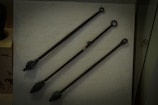 Three elongated metal objects with ring-like handles displayed on a textured background. Each object has a distinct end, suggesting they could be medieval or ancient tools or weapons.