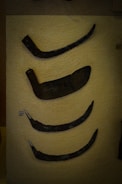 A display showcases a collection of ancient metal tools or weapons, each with a distinct curved shape. They rest against a textured, beige background with small labels indicating numbers for identification. The artifacts appear aged, with a dark, weathered patina.