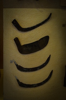 A display showcases a collection of ancient metal tools or weapons, each with a distinct curved shape. They rest against a textured, beige background with small labels indicating numbers for identification. The artifacts appear aged, with a dark, weathered patina.