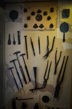 A display case contains an assortment of historical metal tools and objects, including pliers, hammers, and decorative items, arranged on a textured background.