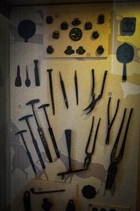 A display case contains an assortment of historical metal tools and objects, including pliers, hammers, and decorative items, arranged on a textured background.