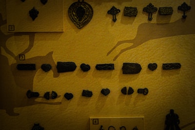 A collection of small, dark metal artifacts are displayed on a textured yellow background. The items include geometric shapes and intricate designs. The display is compartmentalized by small labeled sections, each containing different artifacts.
