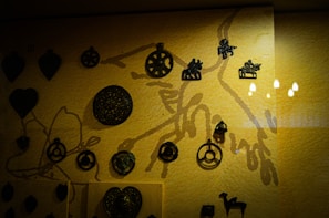 A collection of small, dark metal artifacts displayed against a textured yellow background. The artifacts include circular designs, figurines of people and animals, and heart-shaped objects. The lighting is warm, with reflections visible on the surface.